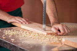  A close-up of someone's hands manually feeding a piece of wood through a bandsaw with wood shavings on the table.